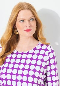 Abito viola con grandi pois bianchi, scollatura rotonda e maniche lunghe. La modella ha capelli ondulati e orecchini a cerchio dorati.