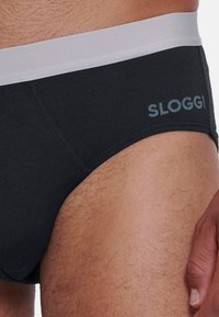 Schwarze Baumwollslips mit einem weißen Gummibund, der das "Sloggi"-Logo in Hellblau zeigt. Strukturiertes Material und glatte Kanten sind sichtbar.