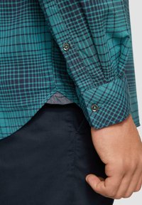 Groene en marineblauwe geblokte shirt met lange mouwen, knoopmanchetten en een gebogen zoom, gemaakt van een gestructureerde stof, die stikdetails benadrukt.