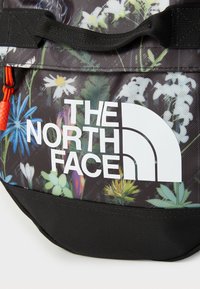 Black fabric backpack with a floral pattern, including blue and white flowers, featuring a white logo and red zipper pull. Textured surface.