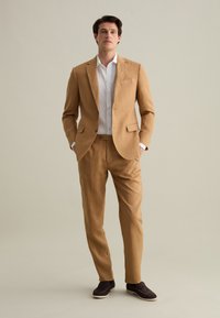 Completo in lino marrone chiaro con giacca a una button e pantaloni a gamba dritta. Abbinato a una camicia bianca con bottoni e scarpe scure.