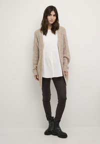 Gilet en tricot beige à front ouvert, porté sur un chemisier blanc et un pantalon noir, associé à des bottines noires à semelles épaisses. Texture lisse.