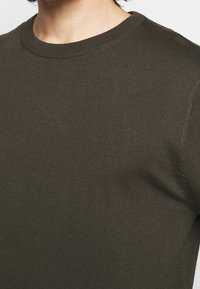 Primo piano di una persona che indossa un maglione a girocollo lavorato a maglia di un verde oliva scuro.