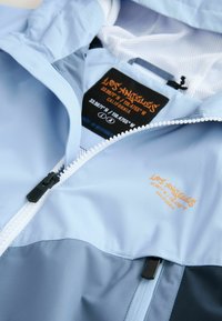 Ljust blå jacka med dragkedja, mesh-foder och svarta detaljer. Har en orange logotyp och koordinater på lappen.