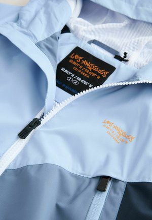 Veste bleu clair avec fermeture zippée, doublure en mesh et accents noirs. Présente un logo orange et des coordonnées sur l'étiquette.
