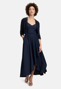 Robe de soirée bleu marine avec une jupe fluide, un corsage ajusté et un décolleté en V, accompagn ée d'un couvre-épaules court et moelleux. Talons argentés.