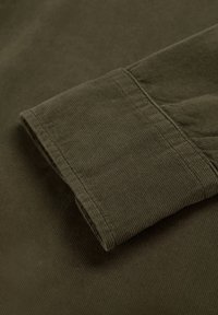 Manche de chemise à col en velours côtelé olive foncé, présentant une texture nervurée et des coutures soignées. Le poignet roulé révèle la finition douce du tissu.