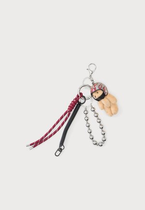 Porte-clés avec un petit ours en peluche beige portant un casque rouge à motif de flammes, attaché à plusieurs anneaux métalliques et cordons rouges et noirs.