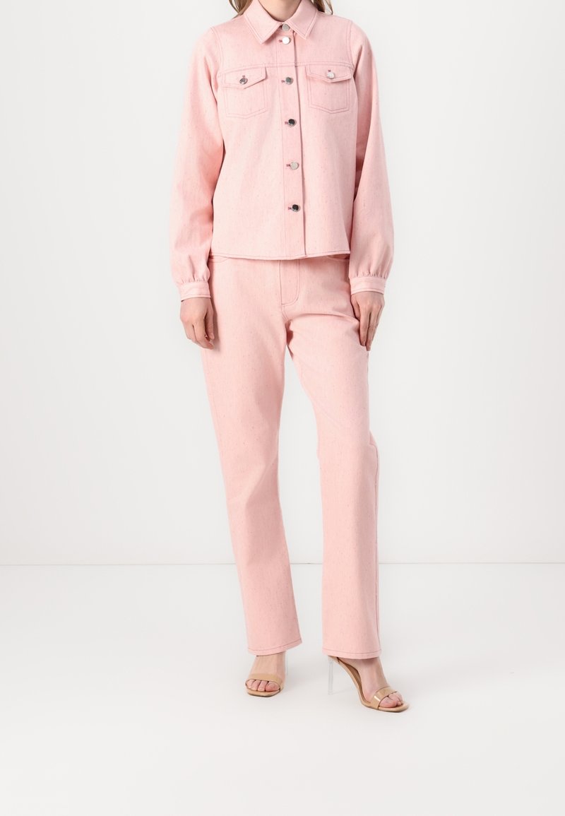 Femme portant une veste en jean rose clair avec boutonnière devant, un jean droit assorti, et des sandales à talons avec bride transparente.
