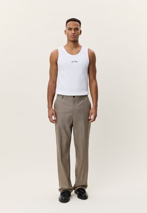 Mees seismas, kandes valget varrukateta särk "les Deux" tekstiga, pruunides ruudulistes pükstes ja musti kingi, ühevärvilise tausta ees.