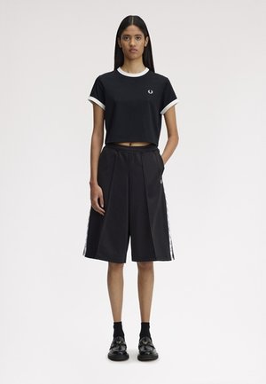 Femme debout portant un t-shirt court noir avec un liseré blanc et un short large noir au genou avec des bandes latérales.