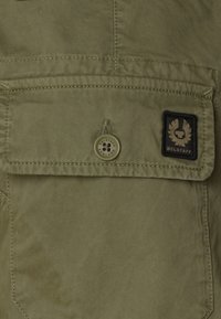 Olive-green fabric shirt with a flap pocket, featuring a button closure and a black patch logo labeled "Belstaff." Textured cotton material.