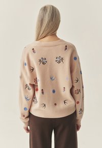 Hele roosa lahtise lõikega kudumistega kampsun, millel on värvikad tikandmotiivid lillede, lindude ja nukude kujul üle kogu pinna.