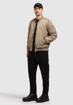 Beżowa kurtka bombka z prążkowanymi mankietami, kieszenią z zamkiem błyskawicznym na rękawie, w połączeniu z czarnymi spodniami cargo i czarnymi, sznurowanymi butami.