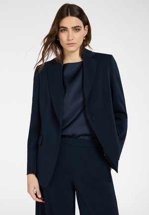 Frau mit langen braunen Haaren, die eine dunkelblaue Anzugjacke und passende Hosen über einer seidigen dunkelblauen Bluse trägt, steht vor einem einfarbigen Hintergrund.