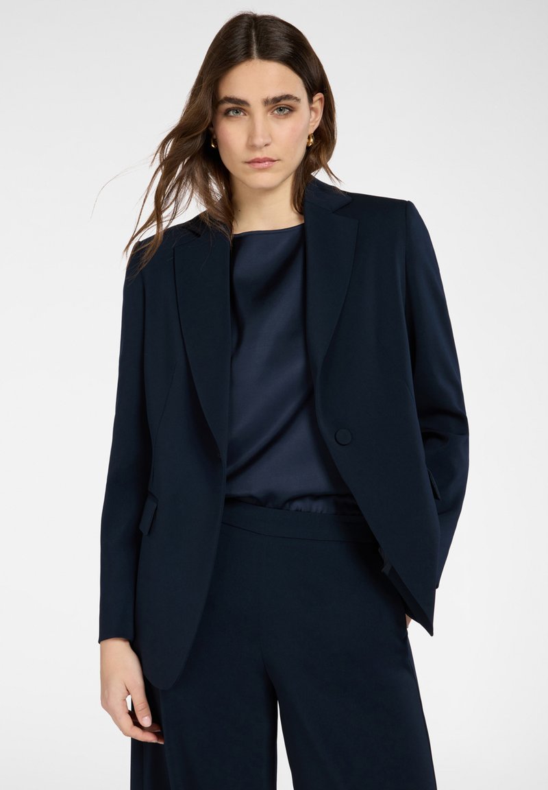 Frau mit langen braunen Haaren, die eine dunkelblaue Anzugjacke und passende Hosen über einer seidigen dunkelblauen Bluse trägt, steht vor einem einfarbigen Hintergrund.