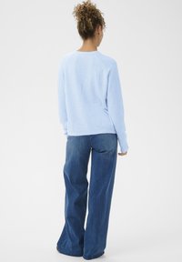 Göndör hajú nő világoskék pulóvert és bő szárú kék farmert visel, háttal áll egy fehér háttér előtt.