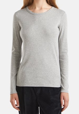 Vrouw draagt een eenvoudige lichtgrijze lange mouwen shirt en zwarte broek, staand tegen een witte achtergrond.