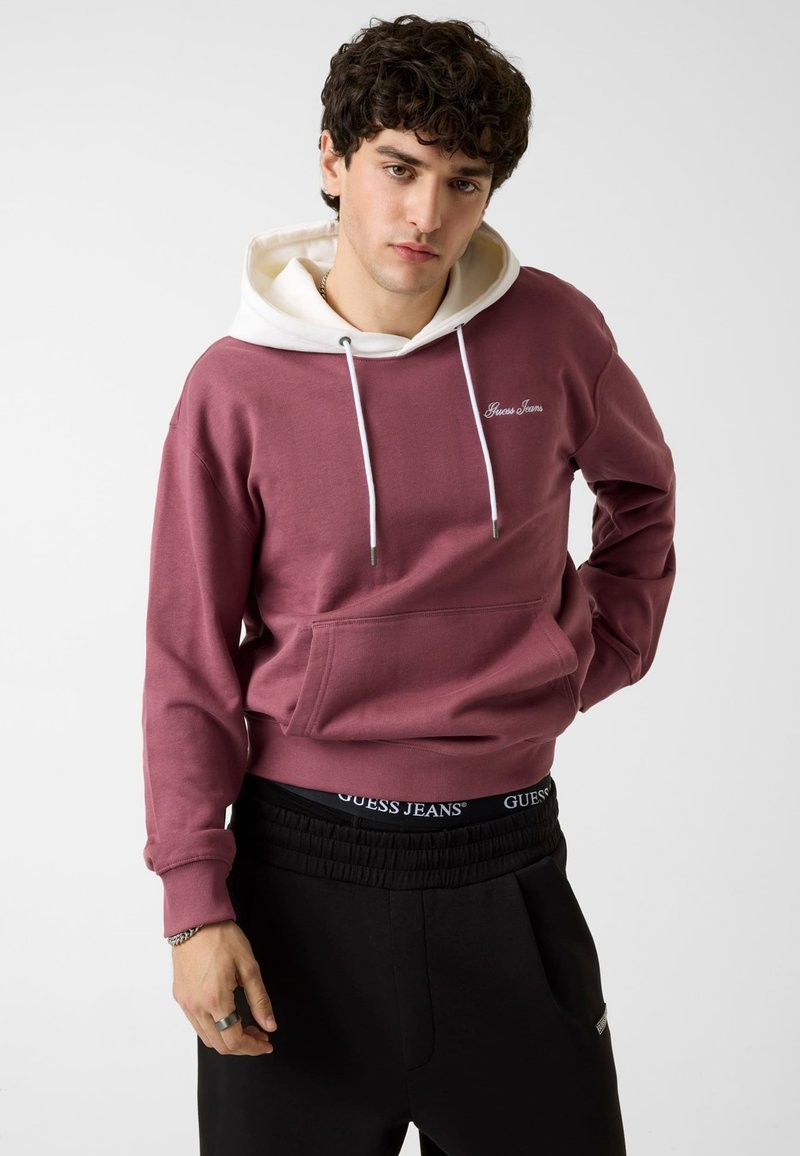 Jeune homme aux cheveux foncés et bouclés porte un sweat à capuche bordeaux avec une capuche blanche et un pantalon noir, debout devant un fond clair et uni.