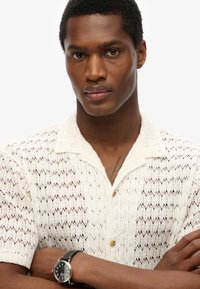 Camisa blanca de punto de manga corta con un patrón de zigzag texturizado y cuello. Llevada con un reloj negro que tiene una esfera redonda y acentos plateados.