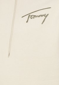 Tissu couleur crème avec un logo "Tommy" brodé gris clair et un cordon crème avec une petite extrémité siglée.