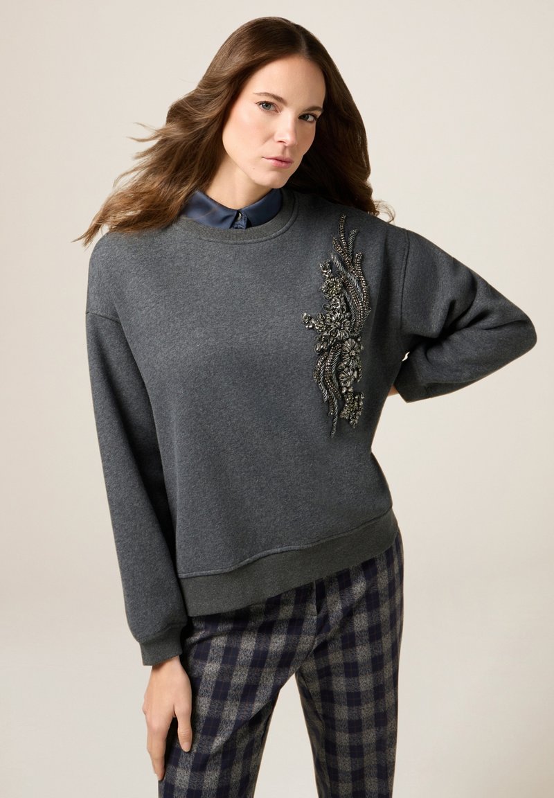 Grauer Hoodie mit einem strukturierten gestickten Design auf der Vorderseite, kombiniert mit karierten Hosen. Verfügt über einen runden Ausschnitt und lange Ärmel.