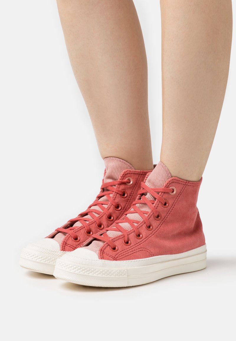 Personne portant des baskets montantes en toile rose avec des semelles en caoutchouc blanc et des chaussettes roses assorties, debout sur une surface blanche.
