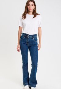 Hochgeschnittene ausgestellte Jeans aus dunkelblauem Denim mit zwei Vorderaschen, kontrastierenden Nähten und einer Knopfleiste, kombiniert mit einem weißen Kurzarmhemd.