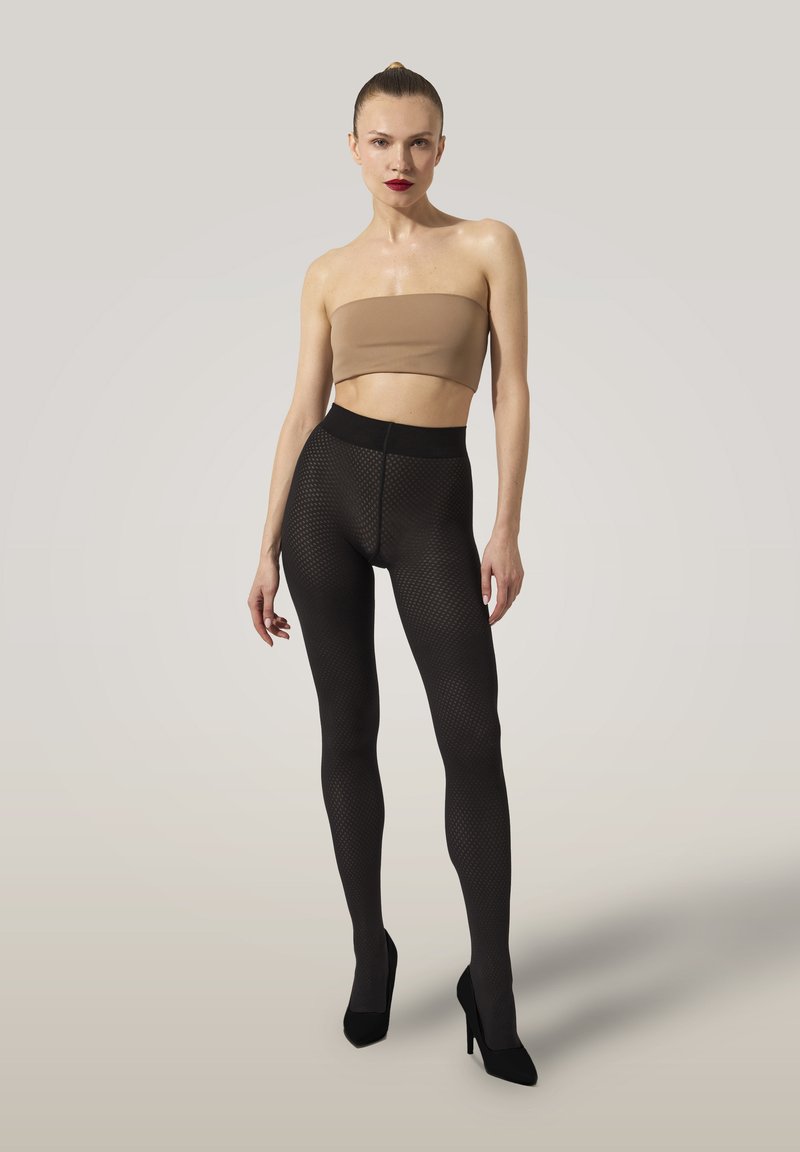 Kvinna som står barfota i klackar och bär en beige strapless topp och svarta texturerade leggings mot en enkel bakgrund.