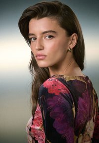 Porträt einer Person mit langem dunklem Haar, die ein Oberteil mit floralem Muster in Rosa- und Lilatönen trägt und kleine goldene Creolenohrringe hat.