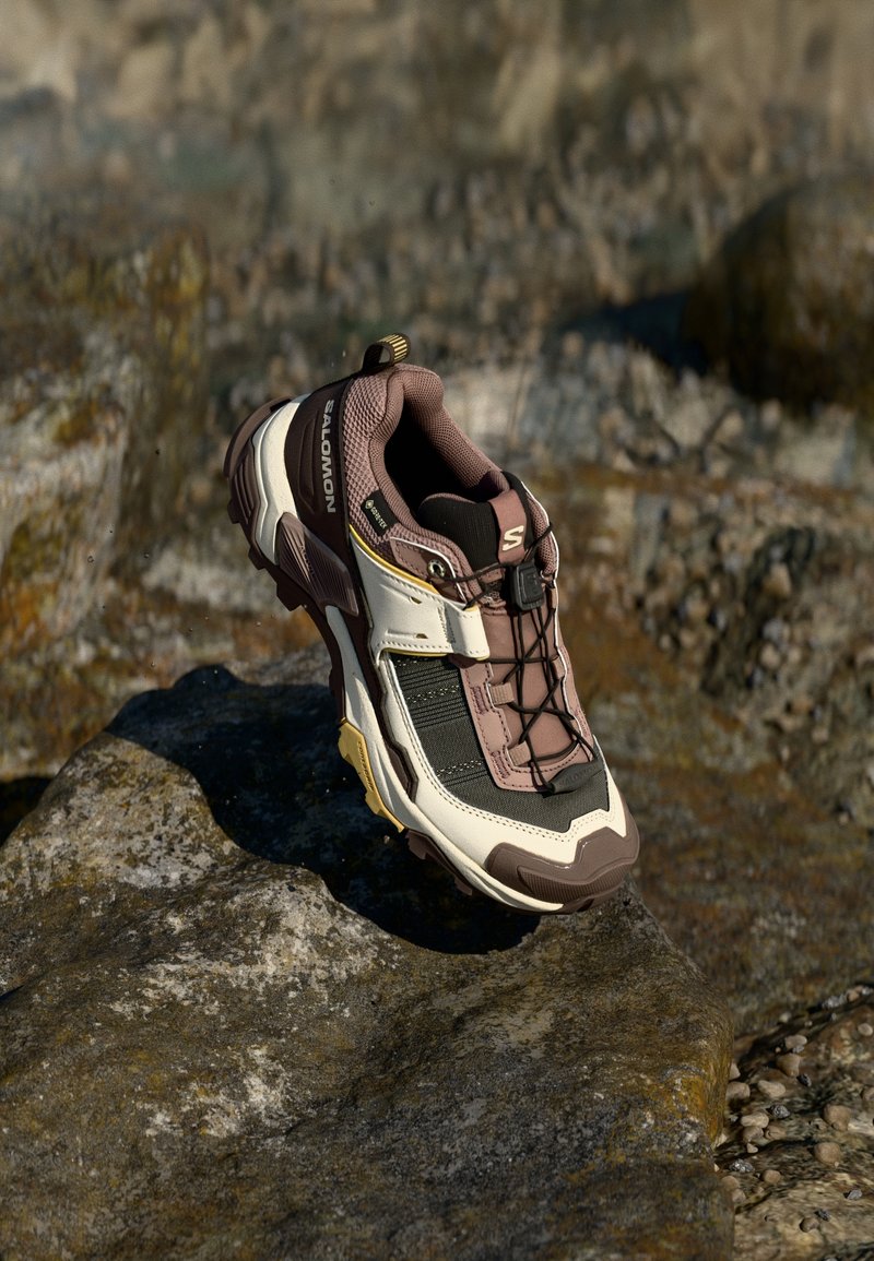Chaussure de trail Salomon en marron, noir et blanc posée sur un gros rocher en extérieur.