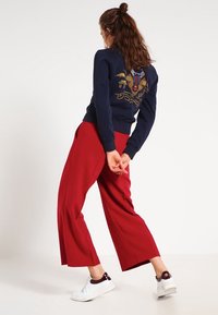 Námořnická mikina s vyšitým designem na zadní straně, doplněná širokými červenými kalhotami a bílými teniskami, které ukazují uvolněný siluetu.