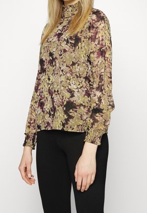 Blouse florale avec un col haut, présentant un mélange de teintes violettes et vertes, manches longues avec des poignets élastiques, tissu doucement texturé.