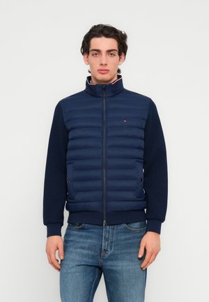 Marineblå lynlåstrøje med polstret front, ribstrikkede ærmeafslutninger og nederste kant. Har et lille logo på brystet og en mock neck-design.