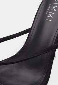 Μαύρο δερμάτινο σανδάλι με λεπτές μαύρες λουρίδες και ορατές ραφές κατά μήκος των άκρων, το οποίο είναι τοποθετημένο σε ελαφρώς λοξή θέση πάνω σε λευκό φόντο.