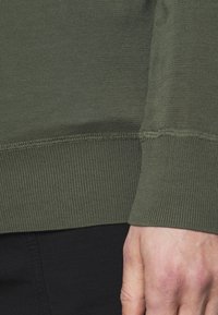 Oliwkowa koszula z długim rękawem, z prążkowanymi mankietami, gładką fakturą i dopasowanym krojem, częściowo włożona w czarne spodnie.