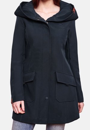 Vrouw draagt een zwarte jas met capuchon, voorvakken met kleppen en een rits die bedekt is met een knoopsluiting, staande tegen een effen achtergrond.