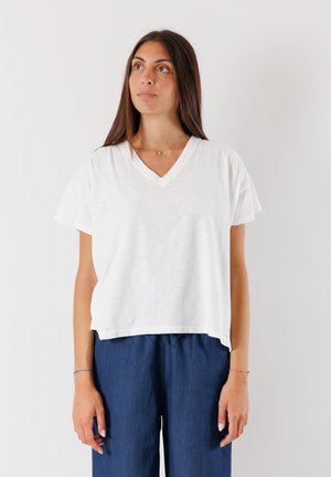 T-shirt bianco a maniche corte con scollatura a V, realizzato in tessuto morbido, caratterizzato da una vestibilità comoda e una leggera texture. Indossato con pantaloni a gamba larga blu.