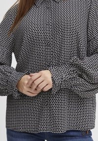 Blusa a pattern bianco e nero con colletto, chiusura a bottoni e polsini arricciati. Realizzata in tessuto leggero con un design fluido.