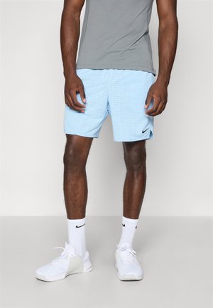 Mann trägt hellblaue Nike-Shorts, weiße Nike-Kniestrümpfe, weiße Sportschuhe und ein graues kurzärmeliges Hemd, steht auf einem grauen Boden.
