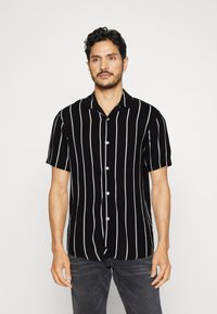 Chemise noire à manches courtes avec boutons, ornée de larges rayures verticales blanches, dotée d’un col classique et d’un tissu léger.