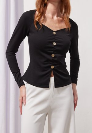 Femme portant un haut noir à manches longues avec des boutons dorés sur le devant et un pantalon blanc taille haute, debout les mains près des hanches.
