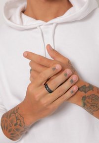 Sort, tekstureret ring på fingrene med forskellige tatoveringer. Hænderne er placeret sammen og viser hudfarve samt detaljer om ringens design.