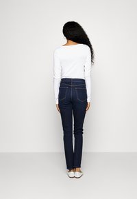 Jeans de mezclilla azul oscuro con cintura alta, que cuentan con dos bolsillos traseros y detalles de costura sutil, combinados con una blusa blanca de manga larga.