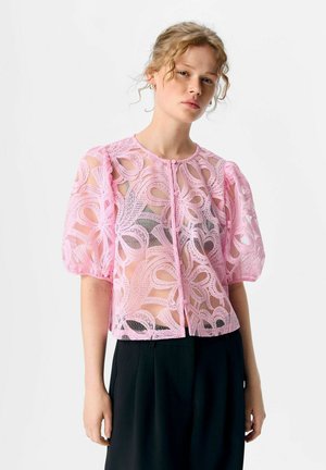 Woman with curly hair wears a sheer pink lace blouse with puff sleeves and black pants against a plain white background.