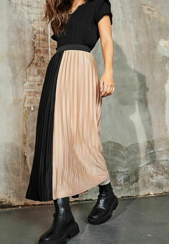 Femme portant un haut noir et une jupe midi plissée avec des blocs de couleur beige et noir, associée à des bottes noires épaisses, debout à l'intérieur.