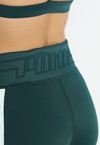 Leggings sportivi verde scuro con una vita alta testurizzata, caratterizzati dal logo "PUMA" in rilievo e da un accento laterale di punti in rilievo per un tocco di eleganza.
