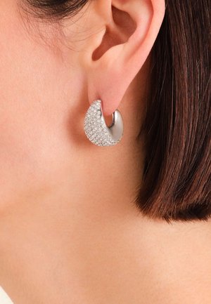 Srebrna naušnica u obliku obruča s polumjesecom u skulpturalnom obliku, s teksturiranom površinom ukrašenom malim prozirnim kristalima.