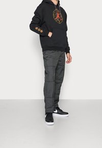 Felpa nera con design geometrico rosso e loghi sulle maniche, abbinata a pantaloni grigi con trama e sneakers nere.