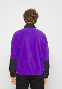 Lilla fleecejakke med sorte ærmer, der har en høj krave og elastisk talje. Blødt materiale, afslappet pasform med korte ærmer.